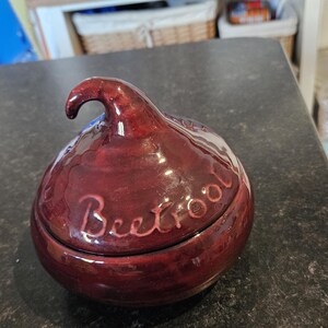 May include: A dark red ceramic container shaped like a beetroot with a lid. The lid has a small handle and the word "Beetroot" is written on the top.