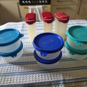 May include: A collection of Tupperware food storage containers including three tall rectangular canisters with red lids and six round bowls with blue, dark blue, and teal lids. The containers are translucent white plastic with matching lids.