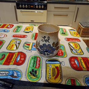 May include: A ceramic salt shaker with a blue floral design and the word "Sel" written on it. The shaker is sitting on a tablecloth with a pattern of colourful sardine cans.