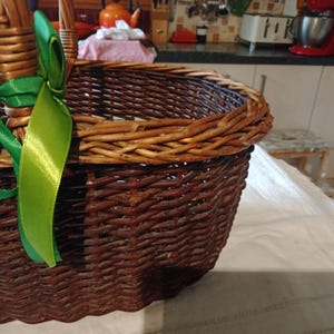 以下が含まれることがあります： 茶色の柳のバスケット。緑色のサテンリボンが結ばれている。バスケットは編み込まれたデザイン。背景にはキッチンと家電製品が写っています。