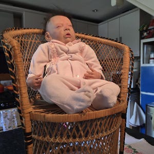 May include: A realistic baby doll with sparse dark hair and closed eyes, wearing a light pink long-sleeved onesie with button closures, seated upright in a tall, woven wicker chair featuring a natural tan finish and a crisscross lattice pattern.