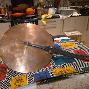 May include: A large, round copper pot lid with a long, black handle. The lid is resting on a colourful patterned cloth with a black and white chequered border. The kitchen background includes a stove, cabinets, and various kitchenware.
