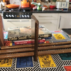May include: A wooden bookend set with two brown bookends. The bookends are holding up a stack of cookbooks, including "Little French Cookbook" and "Delia Smith's Best." The bookends are on a table with a colourful checkered tablecloth.