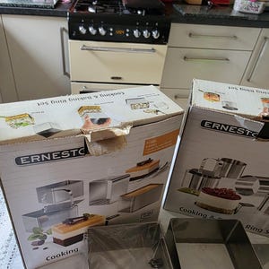 May include: Two boxes labelled "Cooking & Baking Ring Set" and "ERNESTO" are in the foreground. The boxes feature images of cakes and baking tools. A stainless steel square ring is visible in front of the boxes.