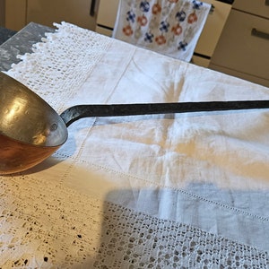 May include: A copper ladle with a long, black metal handle. The ladle is sitting on a white tablecloth with lace trim.