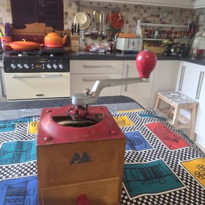 May include: A vintage coffee grinder with a red top and handle, and a wooden base. The grinder has a silver crank and a red knob on the drawer. The grinder is on a patterned tablecloth.