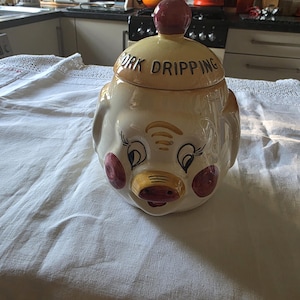 以下が含まれることがあります： ヴィンテージの豚のラードジャー。豚の頭の形をしたジャーで、クリーム色のベース、赤と金の鼻、蓋に赤いノブが付いています。蓋は黄色で、「PORK DRIPPING」の文字が黒で書かれています。