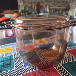 May include: A clear glass casserole dish with a lid. The dish has two handles and is on a colourful tablecloth with a black and white chequered pattern and coloured squares. The dish is likely to hold food.