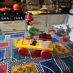 May include: A wooden Pinocchio toy on a yellow cart with red wheels. The toy wears a green jacket, red shorts, and a red hat. The cart has a black and red xylophone and a white container. The toy is on a colourful tablecloth.