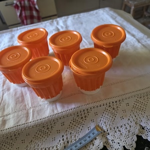May include: A set of six small, fluted containers with bright orange lids and white bases. Each container features a circular, textured pattern on the lid. A blue measuring tape is visible in the foreground, showing a measurement of 20 centimetres.