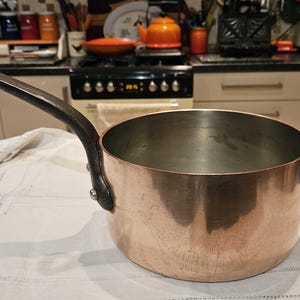 May include: A copper saucepan with a black iron handle. The saucepan is empty and has a shiny, reflective surface.