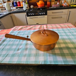 May include: A copper saucepan with a wooden handle and a lid. The saucepan is sitting on a checkered tablecloth.