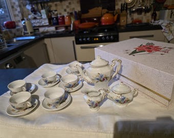 May include: A vintage tea set with a floral design. The set includes a teapot, sugar bowl, creamer, and several teacups and saucers. The pieces are white with blue and pink floral accents. A decorative box is in the background.