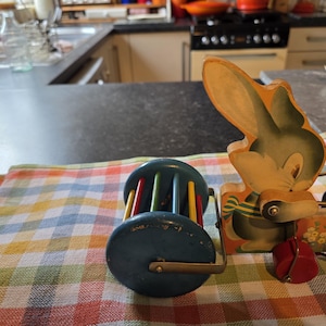 May include: A vintage wooden pull-along toy with a cartoon rabbit design and a rolling drum. The toy is painted in blue, yellow, red, and green. The rabbit has a green and yellow bow tie. The toy is set on a checkered cloth.
