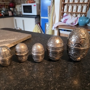 May include: A collection of five silver-coloured, egg-shaped containers, each with detailed embossed patterns. The containers, of different sizes, are arranged in a row on a dark countertop. The background shows a kitchen setting with various items.