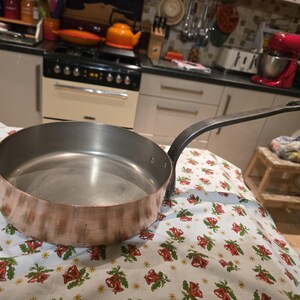 May include: A copper-bottomed sauté pan with a long, dark metal handle. The interior is a light silver colour. The pan sits on a patterned tablecloth with red and green Christmas bell designs. Kitchenware.