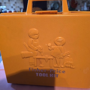 May include: A vintage, mustard-yellow Fisher-Price Tool Kit. The plastic case has a handle and a relief design of two children with tools. The text "Fisher-Price TOOL KIT" is printed below the design.