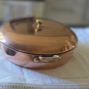 Puede incluir: Una cacerola redonda de cobre con tapa a juego y un pequeño pomo de latón en la parte superior. El recipiente cuenta con dos asas laterales de latón, cada una fijada con dos remaches. La superficie de cobre pulido refleja la luz en su exterior.