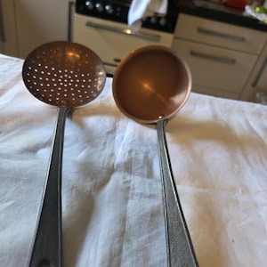 May include: Two copper kitchen utensils: a slotted skimmer and a ladle. Both have copper bowls and grey metal handles with a hole at the end for hanging. The skimmer has many small holes, while the ladle is a solid bowl.