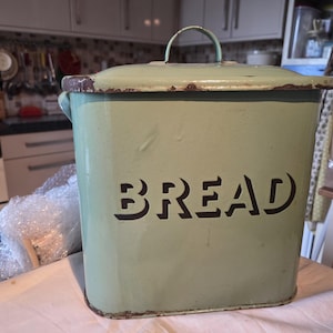 May include: A vintage, rectangular bread bin in a pale green color. The word "BREAD" is cut out on the front. It has a matching lid with a handle and shows signs of wear, giving it a rustic appearance.