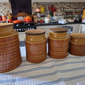 May include: A set of four cylindrical ceramic kitchen canisters with wooden lids, featuring a textured orange and brown floral pattern. The canisters are labeled with embossed text reading BISCUITS, TEA, SUGAR, and COFFEE in a repeating design.