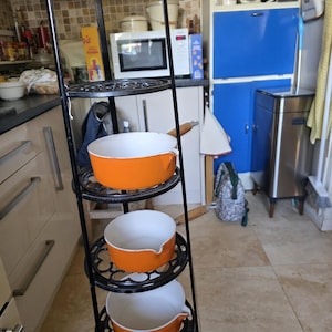 May include: A tall, black metal tiered stand featuring five circular shelves with an intricate openwork pattern. Three orange enamelled metal saucepans with wooden handles are placed on the middle shelves of the vertical storage unit.