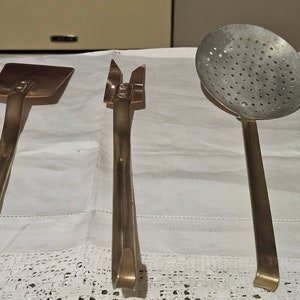 May include: Three vintage kitchen utensils with brass handles. The utensils are a slotted spoon, a spatula, and a pair of tongs.