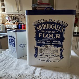 May include: Two white metal storage canisters with blue lids and blue printed text reading McDougall's Self-Raising Flour, featuring a 3 lb net weight label, decorative scrollwork, and the McDougall Bro. signature logo on the front panels.
