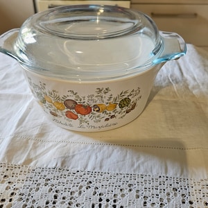 May include: A white glass casserole dish with a clear glass lid. The dish has a floral design with vegetables and the text "Echalote La Marjolaine".