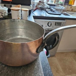 May include: A copper pot with a black handle. The pot is empty and has a shiny, reflective surface.