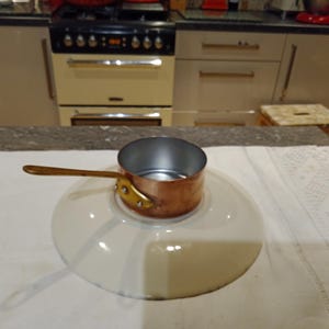 May include: A small copper saucepan with a wooden handle, sitting on a white plate. The saucepan has a shiny interior and is attached to the handle with brass rivets. The plate has a wide rim and a cream colour.