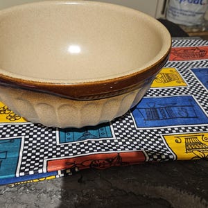 May include: A large, brown and beige ceramic mixing bowl with a fluted rim. The bowl is sitting on a colourful checkered tablecloth.