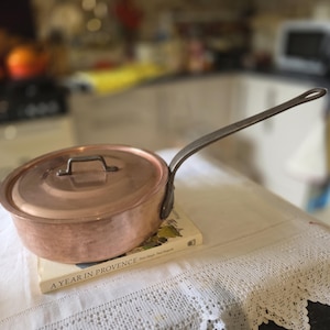 May include: A copper saucepan with a lid and a long, metal handle. The pan rests on a book titled "A Year in Provence" on a white tablecloth. The kitchen background is slightly blurred.