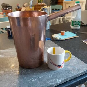 May include: A large copper-coloured metal jug with a long handle and a small white mug with a yellow handle and a colourful design. The jug is tall and cylindrical, and the mug is small and round.