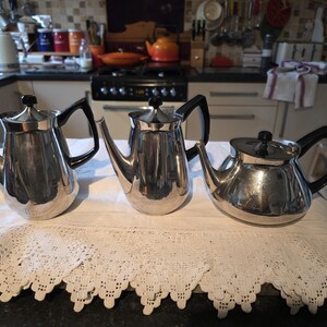 Puede incluir: Tres teteras plateadas con asas y tapas negras dispuestas sobre un mantel de encaje blanco. Las teteras tienen una superficie brillante y reflectante, con un fondo de cocina.