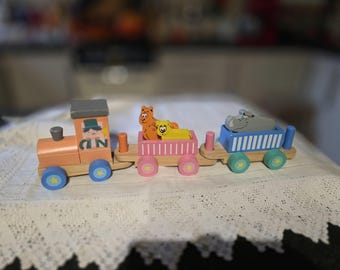 May include: A colorful wooden toy train with three cars. The engine is orange with a gray roof and blue wheels. The first car is pink with two animal figures, and the last car is blue with a hippo figure. The train is on a white surface.