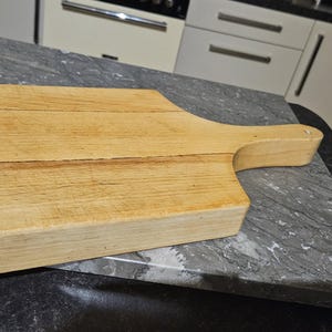 May include: A light brown wooden cutting board with a handle. The board has a rectangular shape and is made of natural wood.