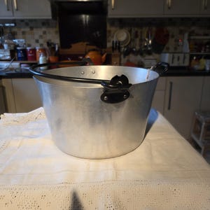 May include: A large, silver metal bucket with a black handle and black side attachments. The bucket sits on a white, lace-edged tablecloth. The background shows a kitchen with cabinets and appliances.