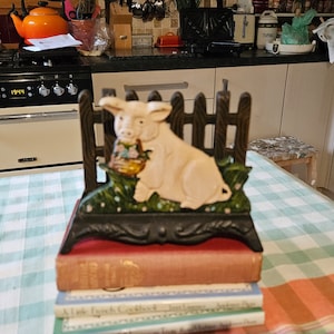 Könnte beinhalten: Ein Napfhalter aus Gusseisen mit einem Schweinedesign. Das Schwein ist weiß mit rosa Akzenten und hält einen Blumenkorb. Der Napfhalter steht auf einem Stapel Bücher.