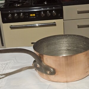 May include: A copper saucepan with a black handle. The saucepan has a shiny copper finish and is sitting on a white tablecloth.