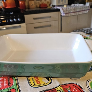May include: A green and white rectangular baking dish with a white interior. The dish is sitting on a table with a patterned tablecloth.
