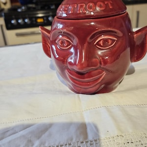 May include: A red ceramic sugar bowl shaped like a face with a wide smile and pointy ears. The lid has the word "Beetroot" written on it.