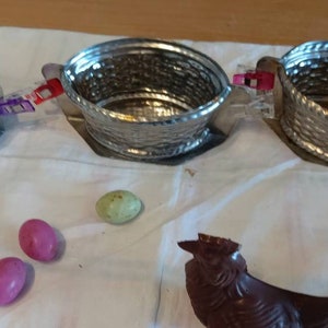 May include: Three silver metal baskets with handles, connected by purple and red clips, on a white cloth surface.  Three chocolate Easter eggs, two pink and one green, and a chocolate Easter bunny are on the surface.