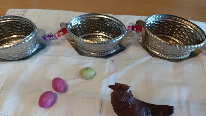 May include: Three silver metal baskets with handles, connected by purple and red clips, on a white cloth surface.  Three chocolate Easter eggs, two pink and one green, and a chocolate Easter bunny are on the surface.