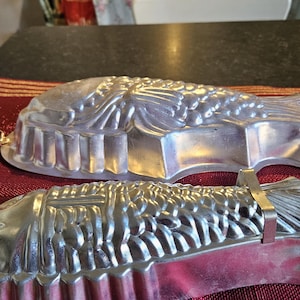 May include: Two silver metal fish-shaped cake moulds. The larger mould is a flat fish with a detailed design. The smaller mould is a more realistic fish with a textured surface.