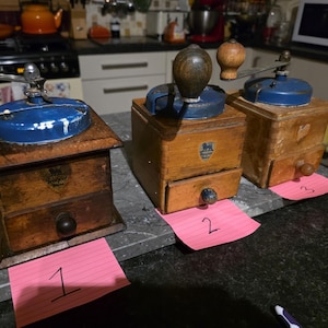 May include: Three antique wooden coffee grinders with blue lids and metal handles. The grinders have wooden boxes with drawers and decorative details. Each grinder sits on a dark countertop, with pink numbered tags in front.