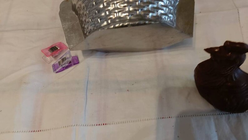 May include: A silver metal basket mold with a woven pattern. A small chocolate bunny figurine is on a white surface.