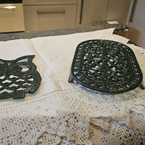 May include: Two green cast iron hot pads, one shaped like an owl and the other with a decorative design, are displayed on a white crocheted tablecloth.