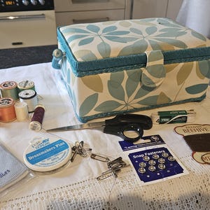 May include: A blue and white floral fabric sewing box with a teal trim. The box is open and filled with various sewing supplies, including spools of thread, needles, pins, and a pair of scissors. There are also several packages of sewing supplies, including snap fasteners, mending fabric, and a package of dressmakers pins.