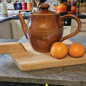 May include: A brown ceramic teapot with a wide spout and a handle. The teapot is sitting on a wooden chopping board with two orange mandarins.